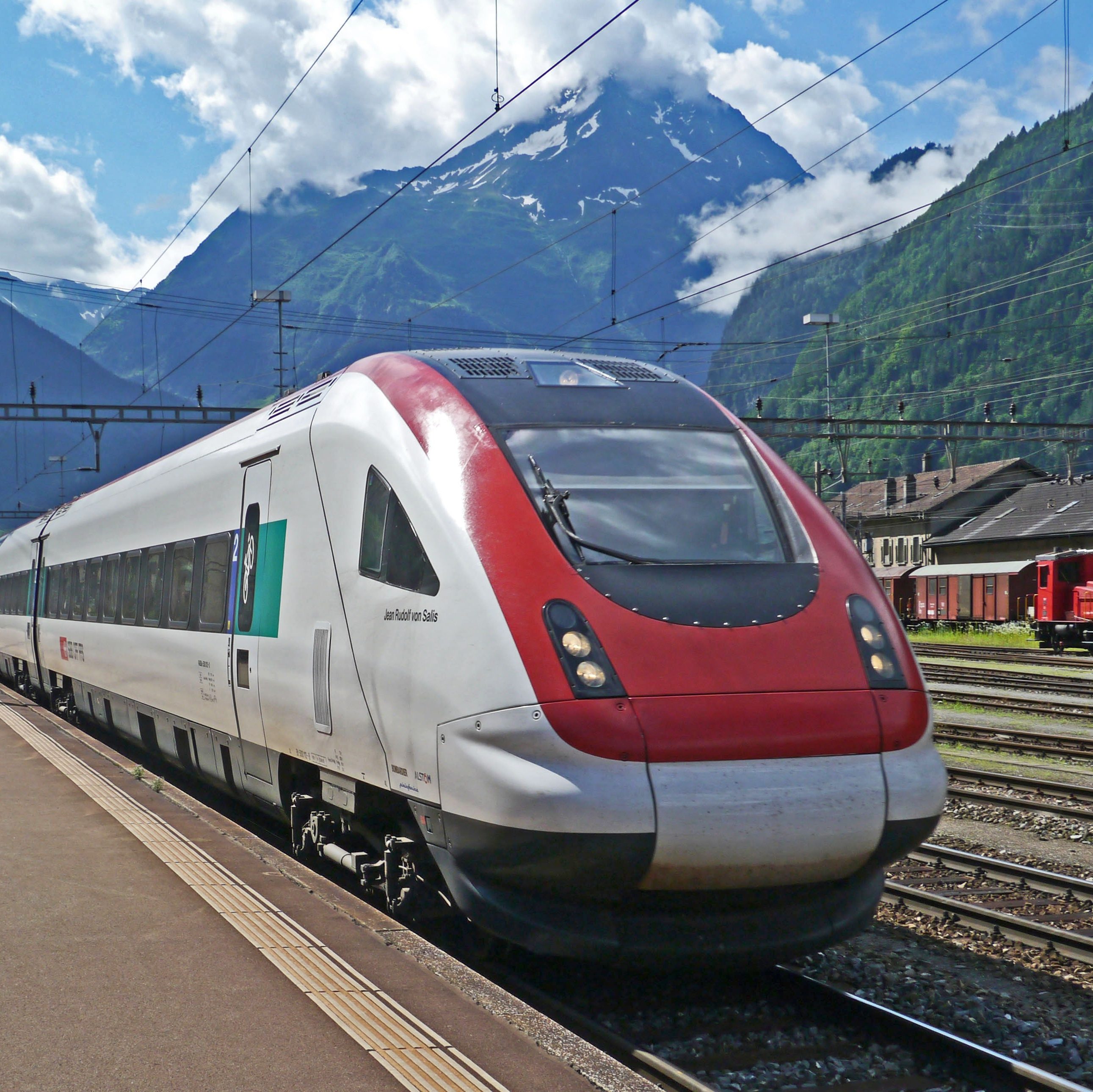 A train is stopped at a station in a mountainous region. Behind the mountains is a cloudy sky.