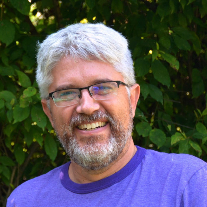 Doug Kaufman smiles at the camera in front of greenery; his short hair and beard are white and he wears rectangle glasses.