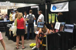 A group of people mill in a convention center. In the foreground are tablets with headphones, and at the local booth we see a banner with the CSCS logo and temperature tapestries.