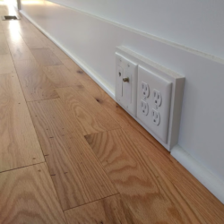 A photo of an electrical wall outlet by the floor, which would be a couple inches away from the wall except that the baseboard reduces the distance to a half inch, and to 0 at the trim.
