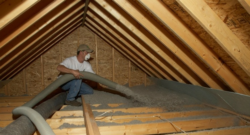 Inside a pitched roof, a man with a face mask blows insulation into the building's rafters.