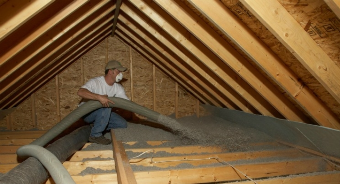 Inside a pitched roof, a man with a face mask blows insulation into the building's rafters.